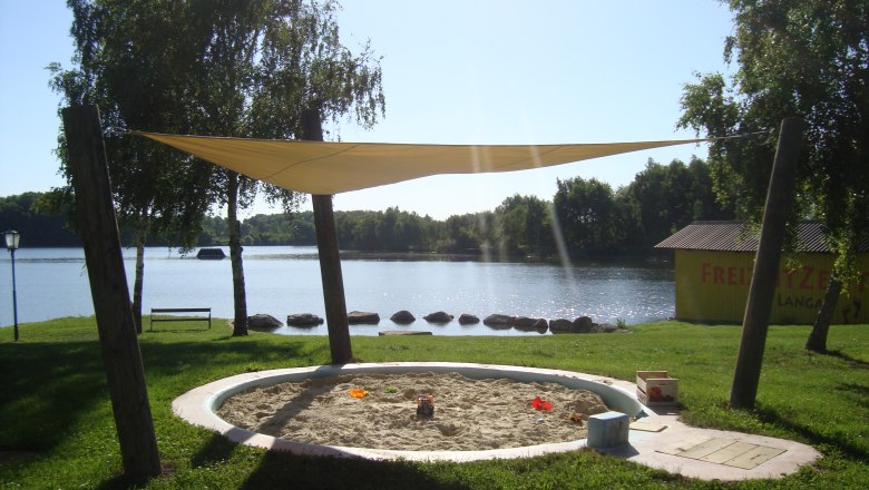 A sandpit with a sun sail on the shore of a lake, surrounded by trees and a meadow. In the background a yellow building with the inscription 'Freizeitzentrum Langau'.