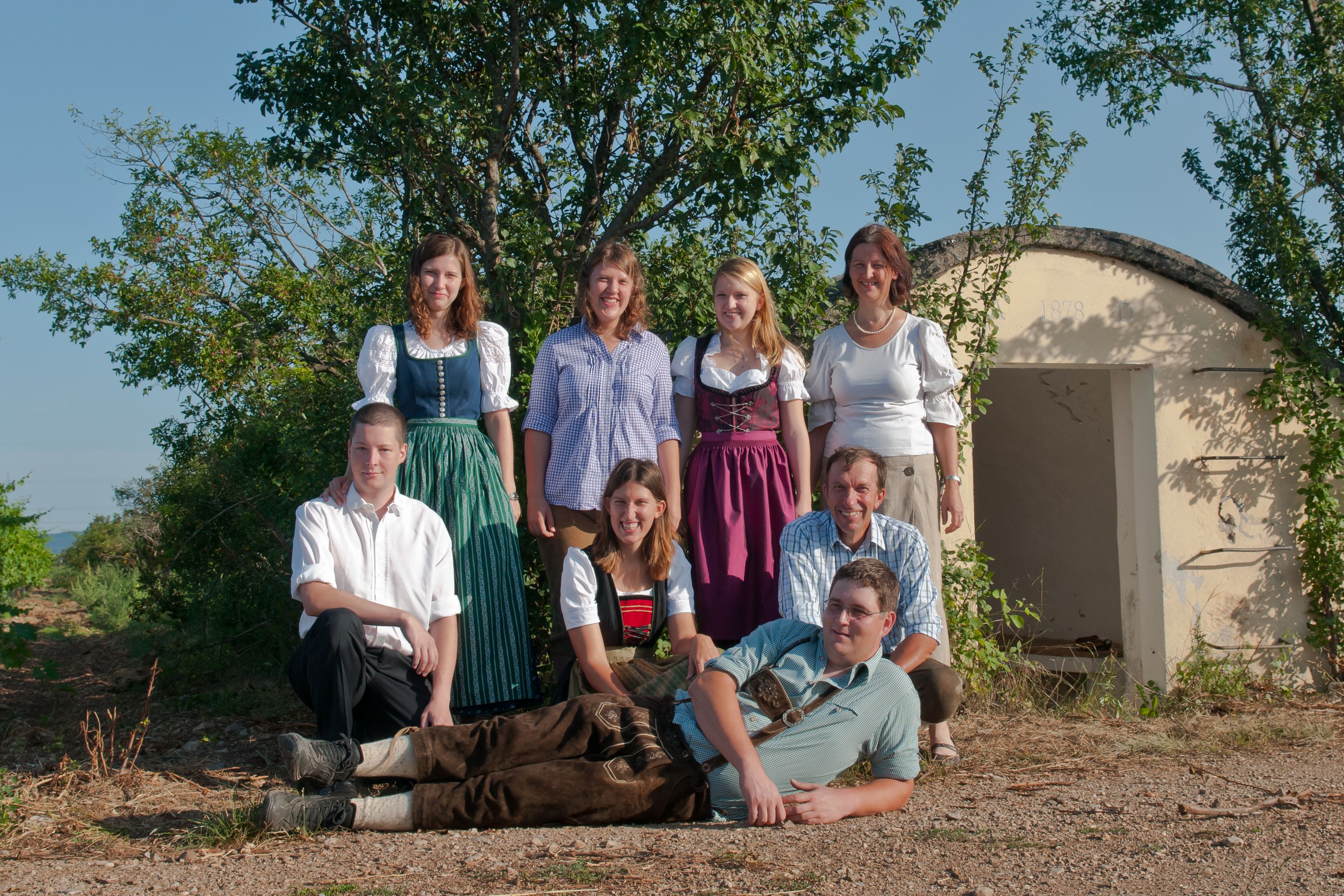 A group of eight people pose outdoors in front of a small building. Some are wearing traditional clothing.