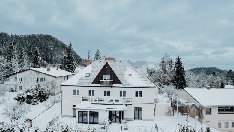 A large, snow-covered house in a wintry landscape with mountains in the background.