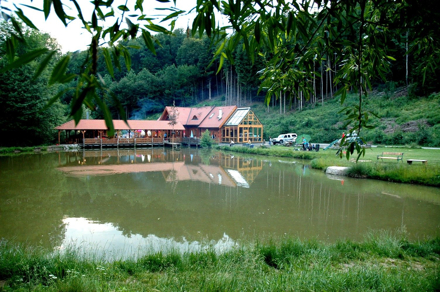 A building with a red roof is reflected in a pond surrounded by trees and meadows.