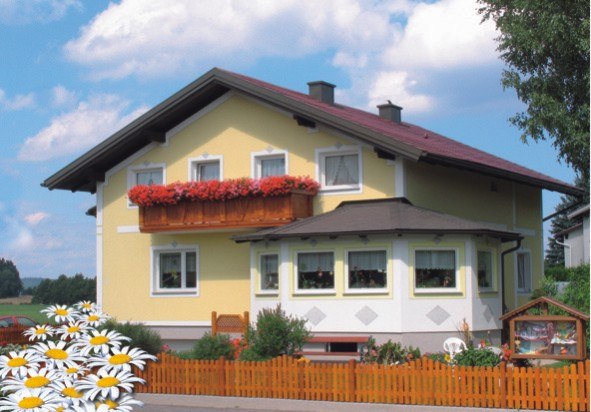 A yellow house with a red roof and a flower box on the balcony, surrounded by a wooden fence and daisies in the foreground.