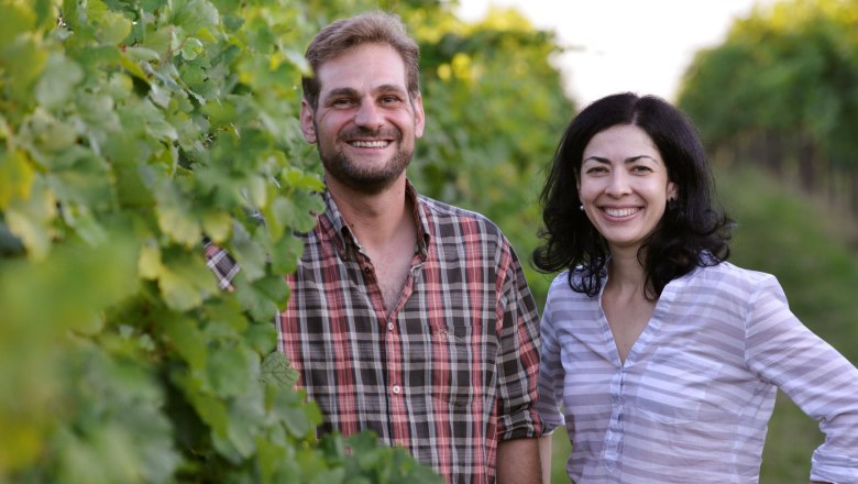 Two people stand smiling in a vineyard.