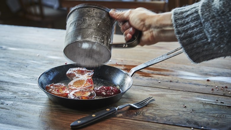 A hand sprinkles powdered sugar over caramelized apple rings in a pan on a wooden table.