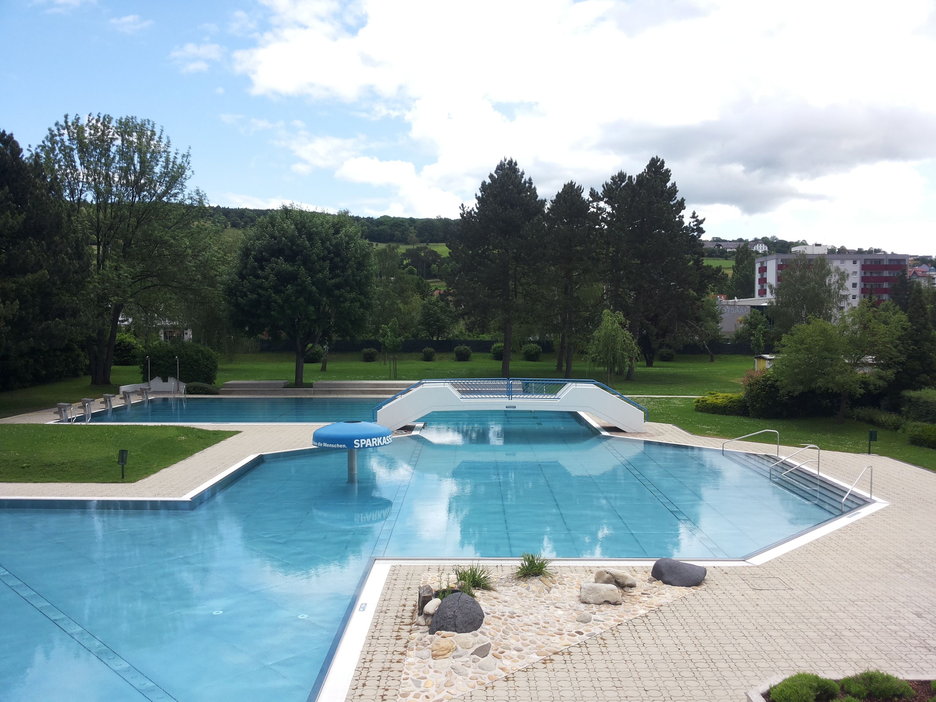An empty outdoor pool with several pools, surrounded by trees and lawns, under a cloudy sky.
