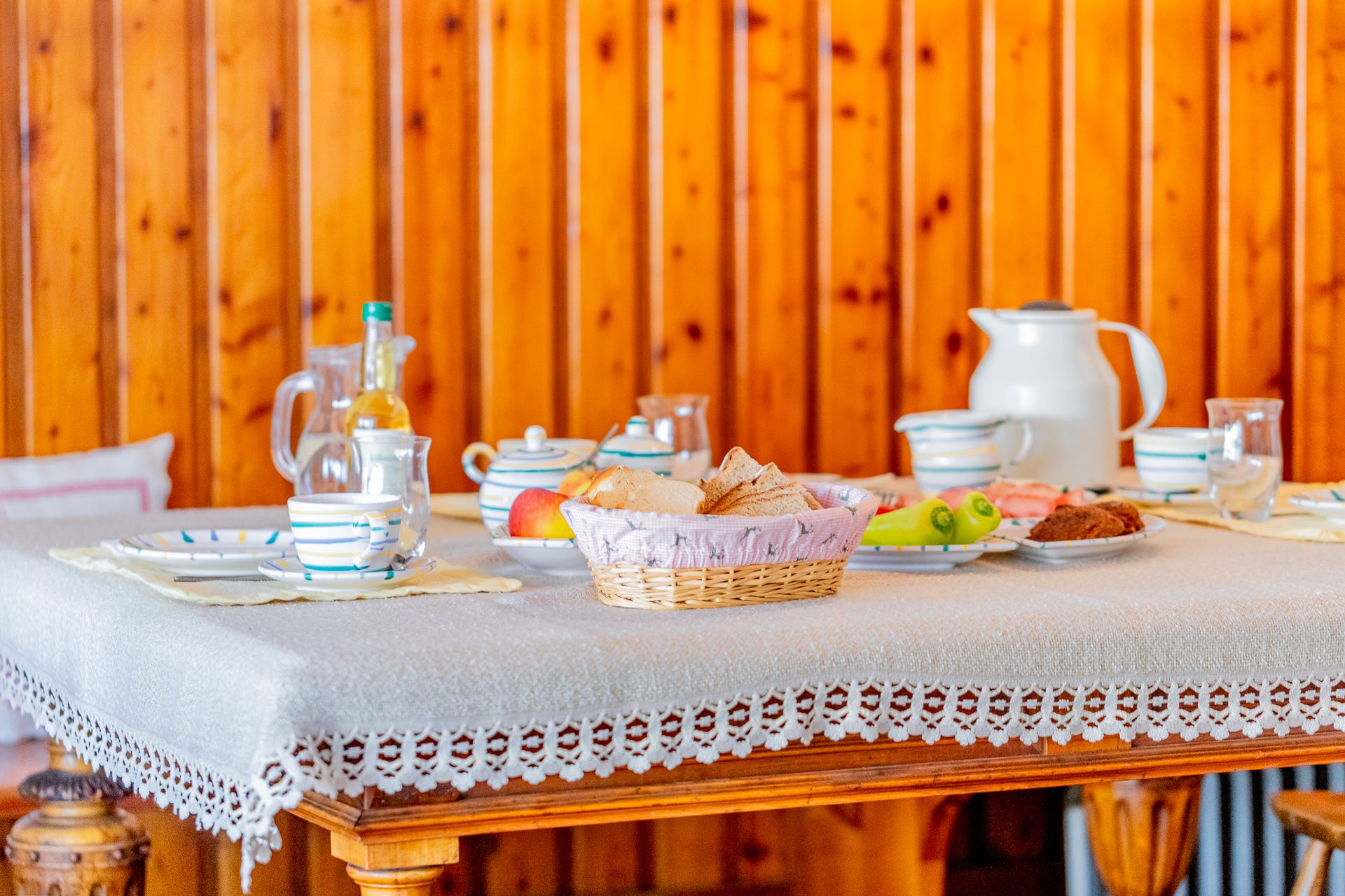 Breakfast table with crockery, bread, fruit and drinks in front of wood paneling.