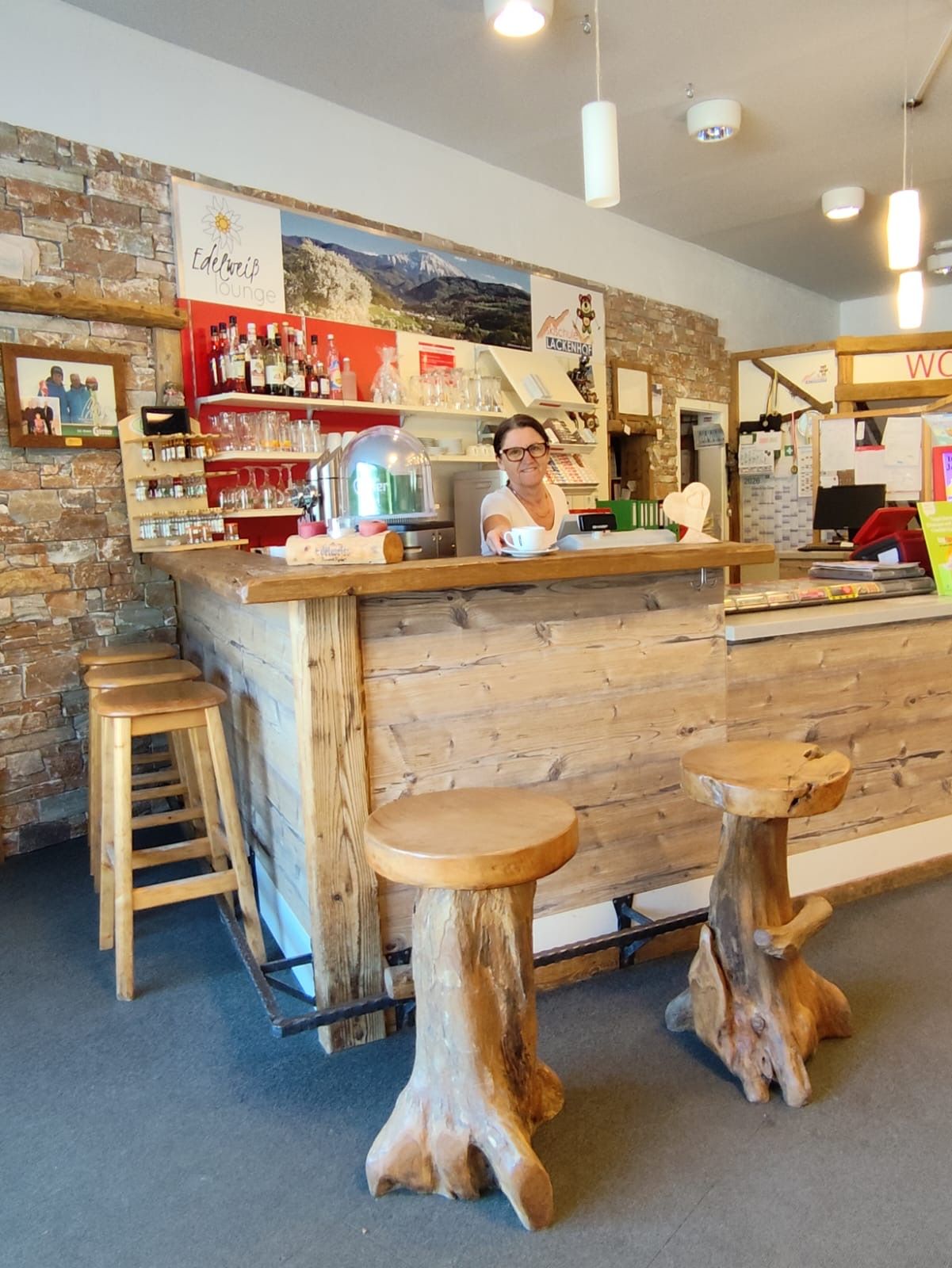 A woman stands behind a rustic wooden counter in a cozy lounge with wooden stools and a stone wall.
