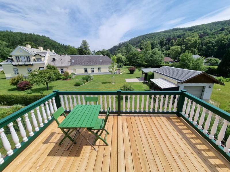 View from a balcony onto a garden with a green meadow and trees, with a large house and hills in the background.