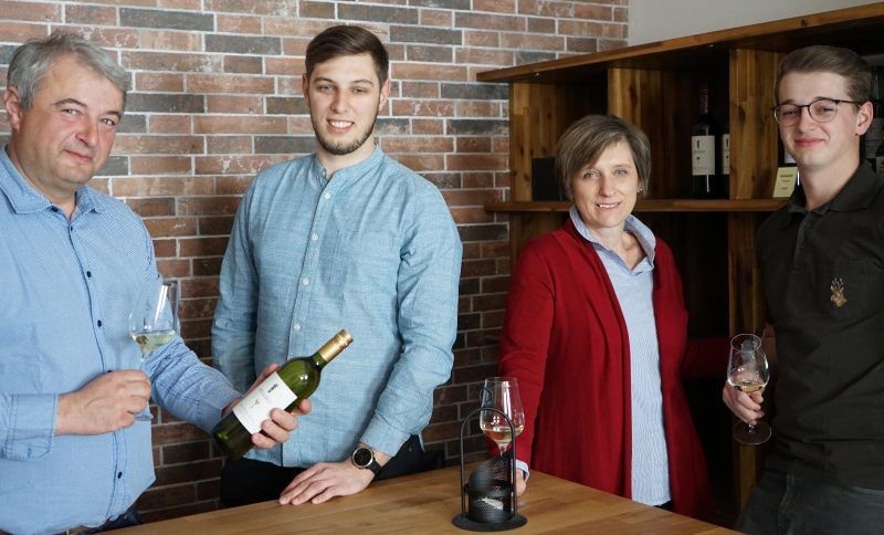 Four people stand at a table with wine glasses and a bottle of wine in front of a brick wall.