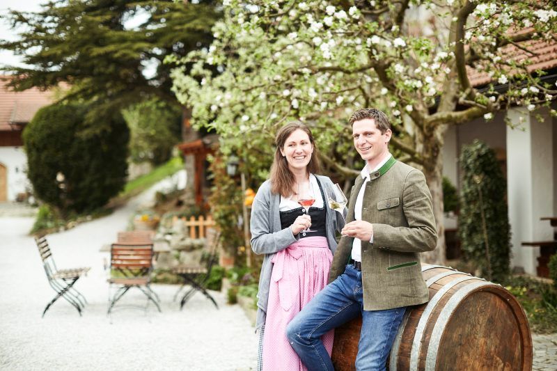 A couple in traditional dress stand smiling with wine glasses in front of a wine barrel outside.