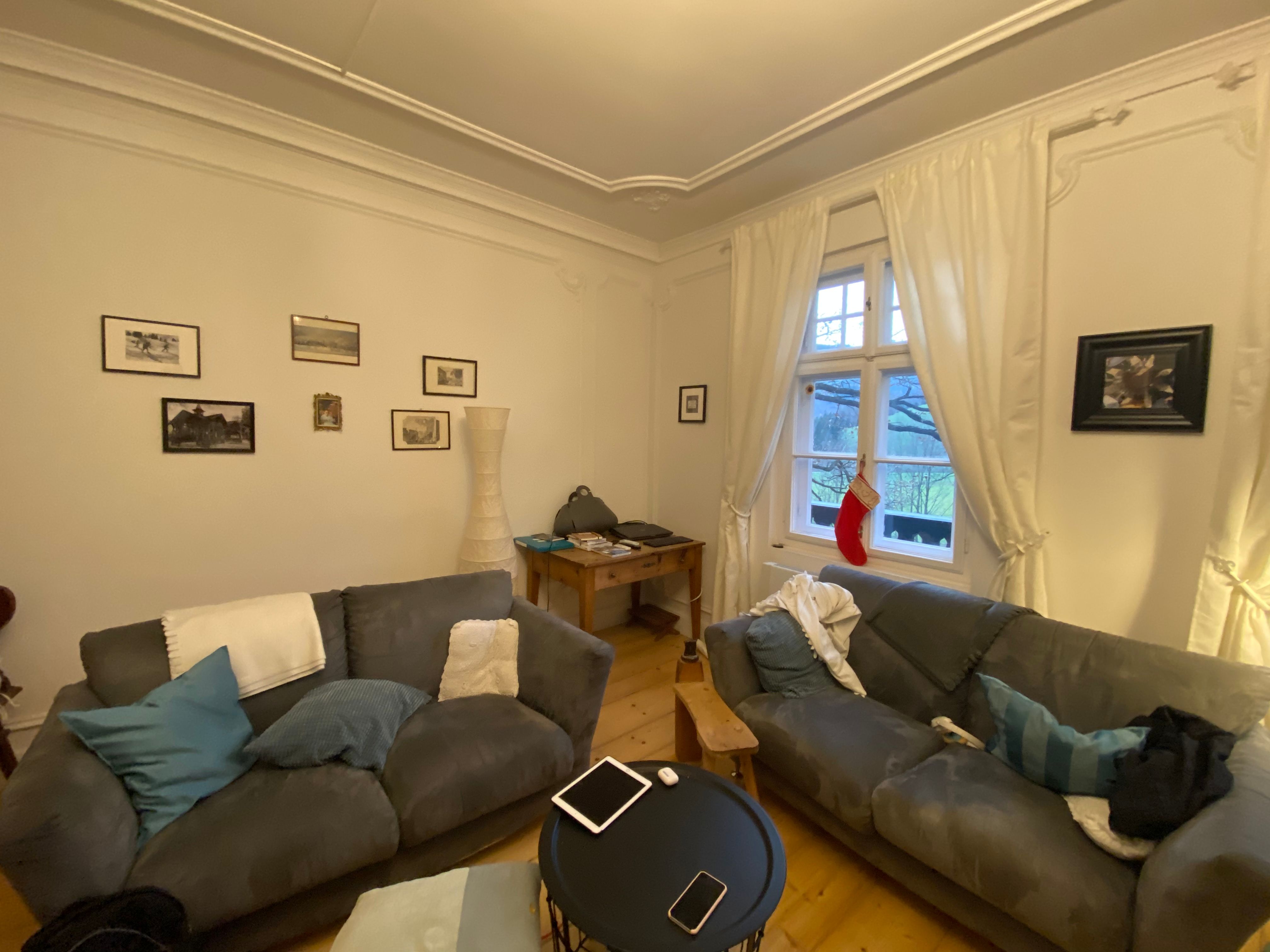 Cozy living room with two grey sofas, a round table, pictures on the wall and a window with curtains and a Christmas stocking.