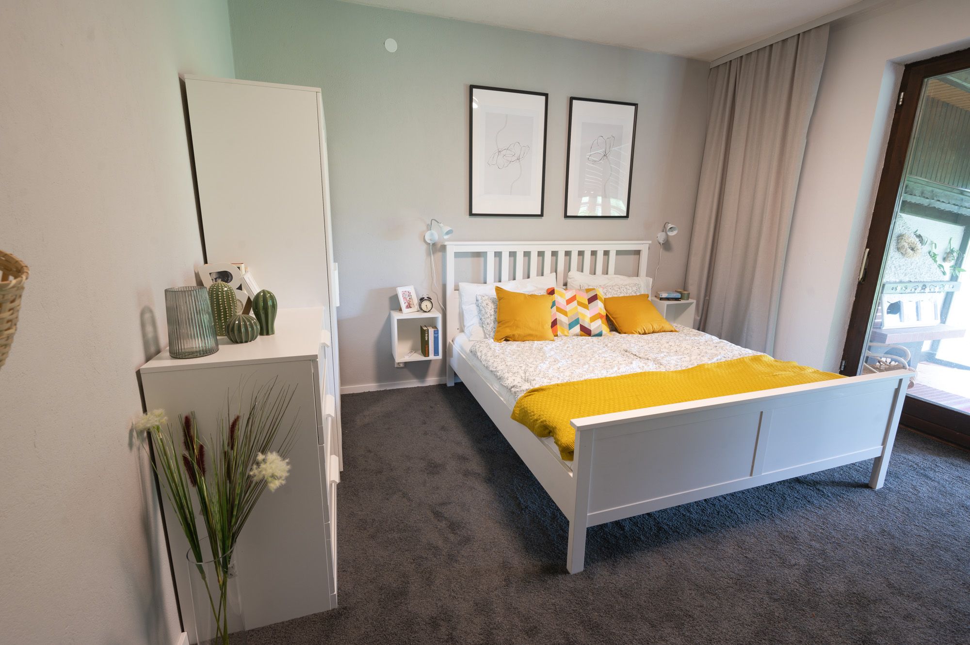Bedroom with white bed, yellow pillows and blanket, two murals and a window with curtain.