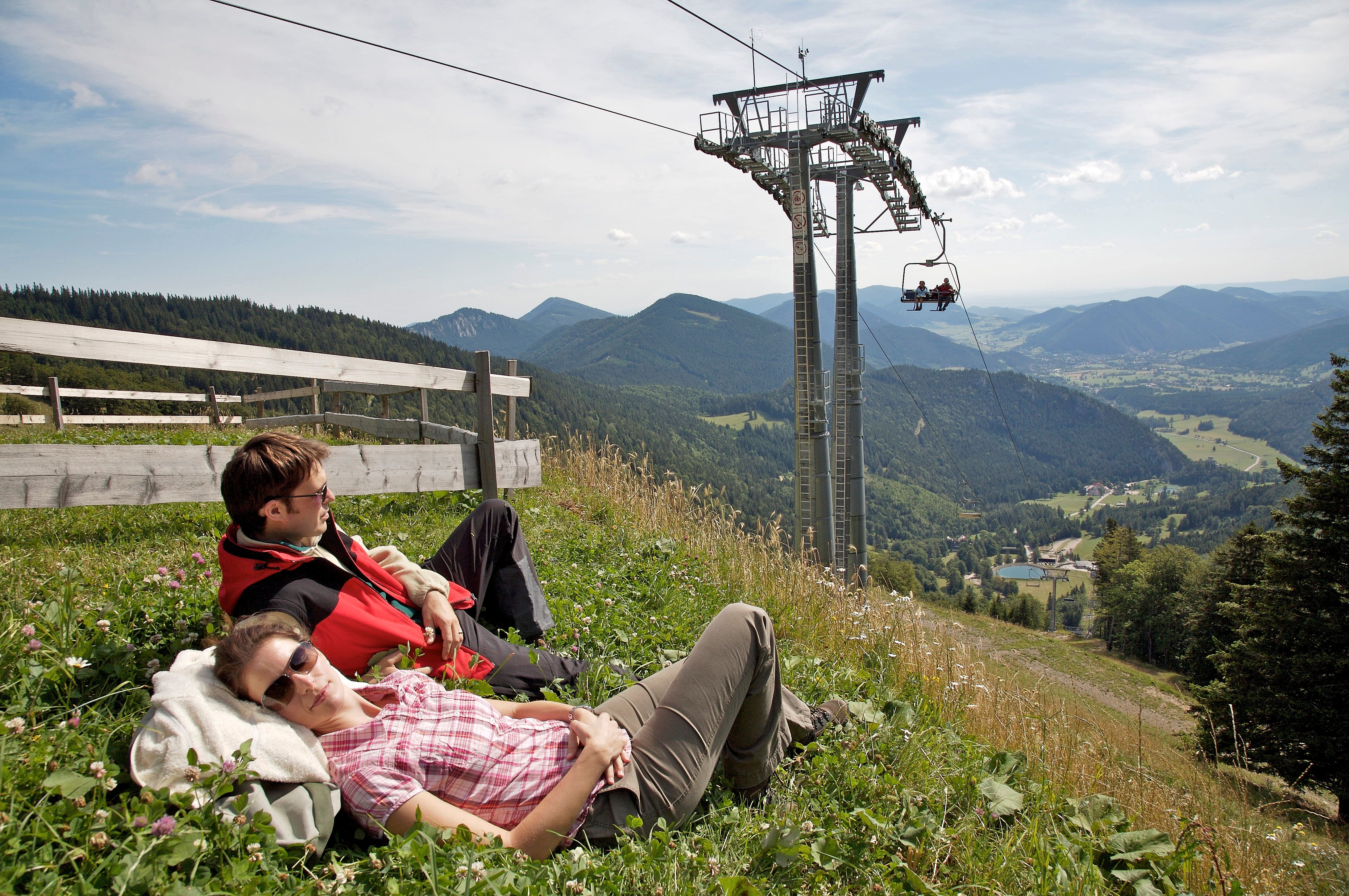 Two people relax on a meadow next to a chairlift with a mountain view.