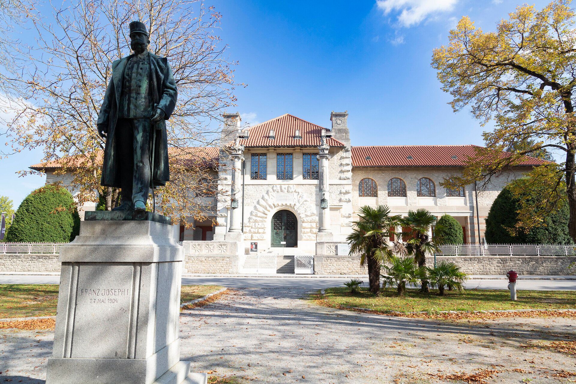 The majestic statue stands proudly in front of an impressive building, surrounded by colorful autumn leaves. The gentle rays of sunlight bathe the scenery in a warm light and invite you to explore the beauty of the surroundings.