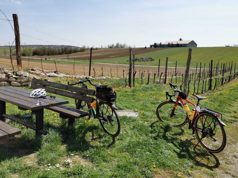 Two bicycles are parked next to a wooden table on a meadow with a view of fields and a building in the background.