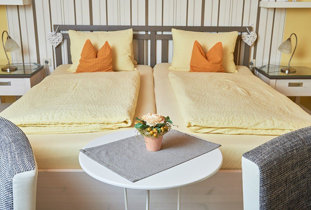 Cozy double room with two single beds, yellow bed linen and orange pillows. A round table with a flower arrangement stands between two armchairs.