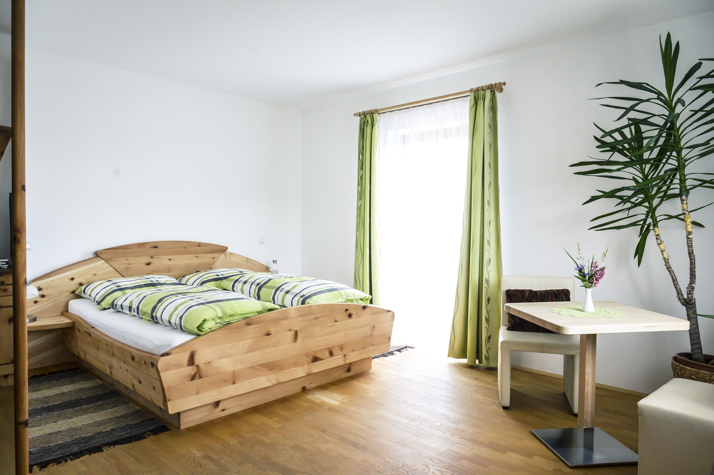 Bright room with wooden bed, green and white striped bed linen, table with vase and plant, green curtains.