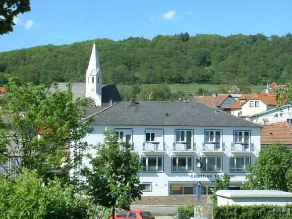 A two-storey building with the inscription 'Gasthof Schauer' in front of a wooded hill and a church with a pointed tower.