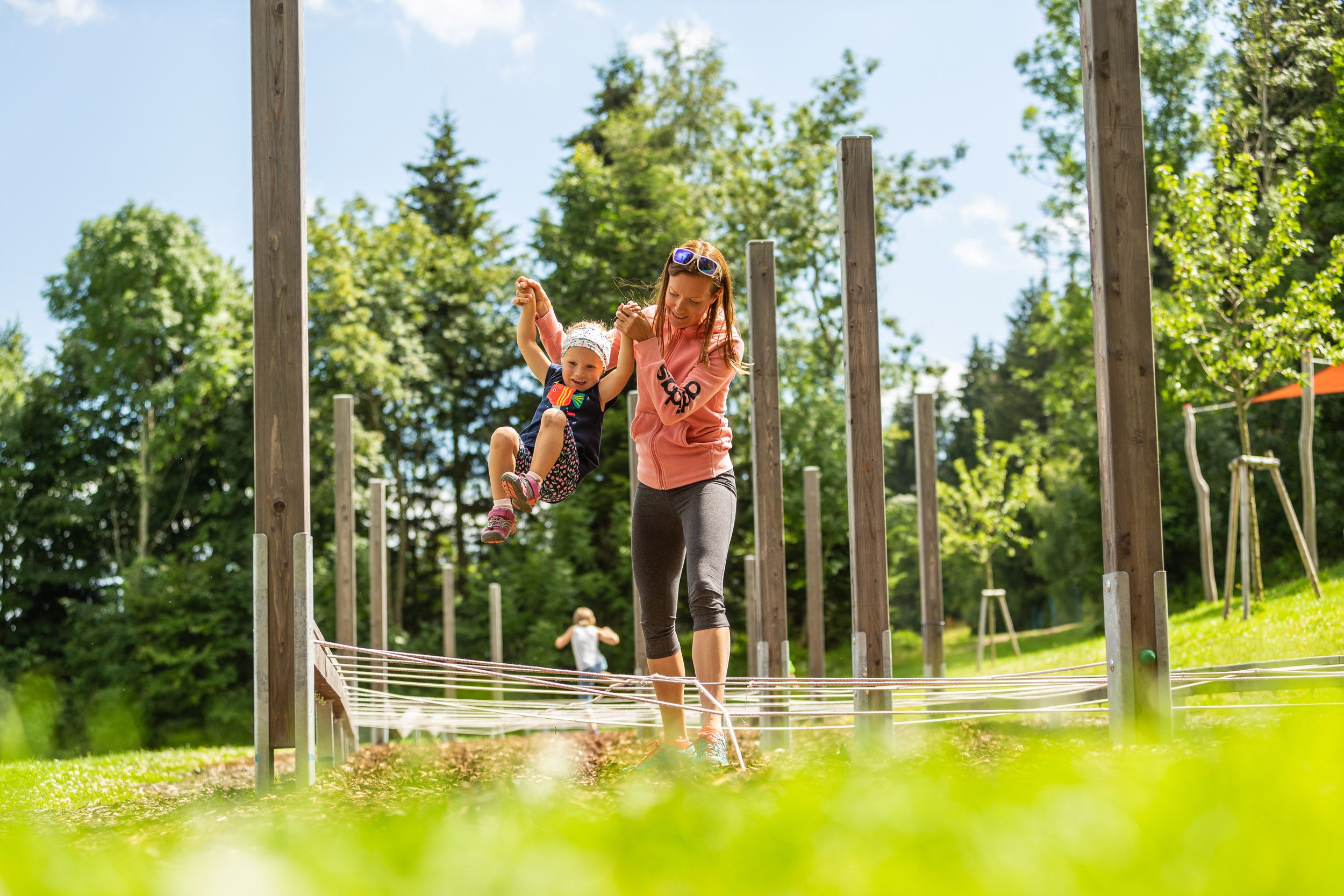 A woman holds a child by the hands in an outdoor play area with wooden posts and ropes.