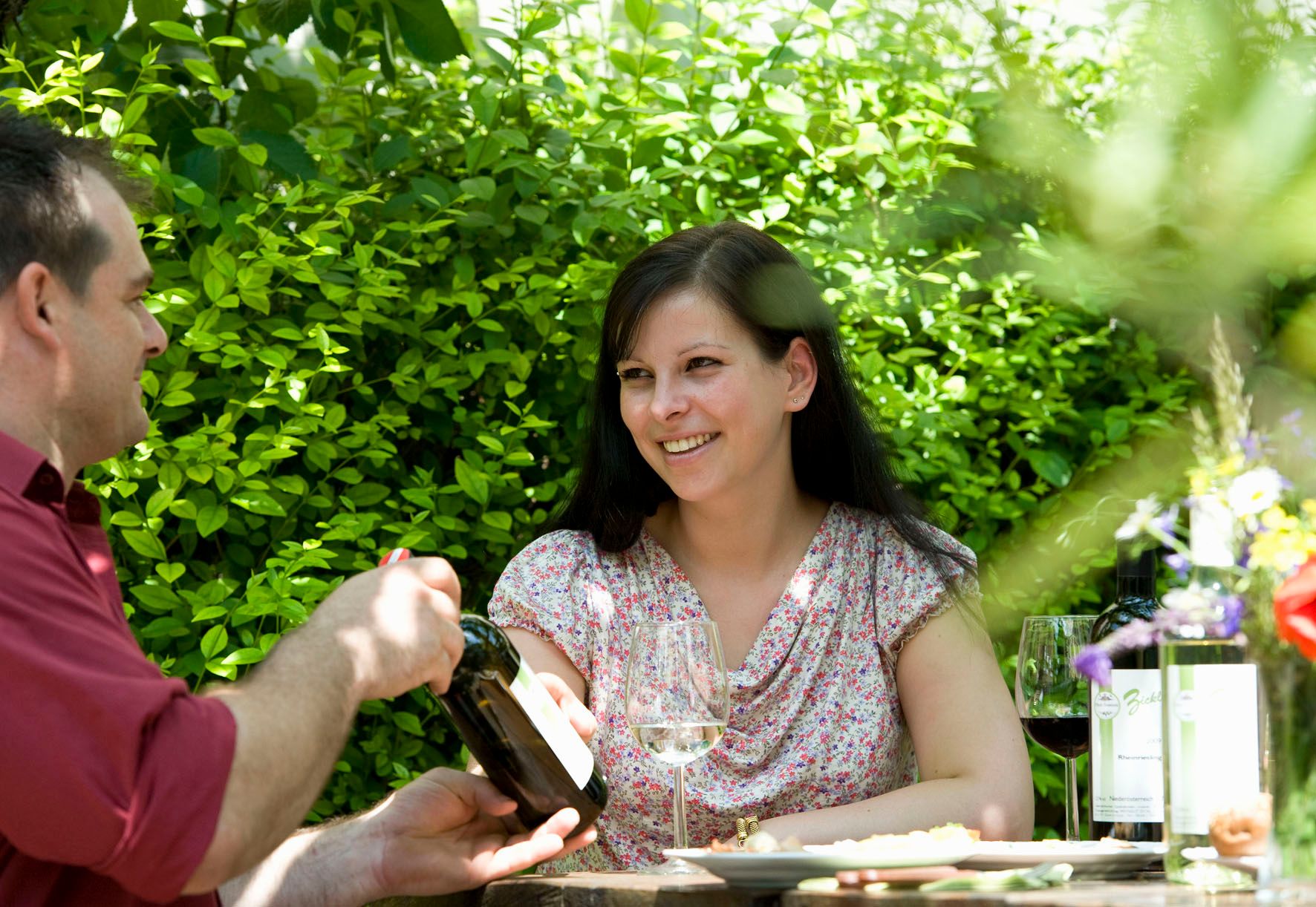 Two people are sitting outside at a table with wine and flowers.