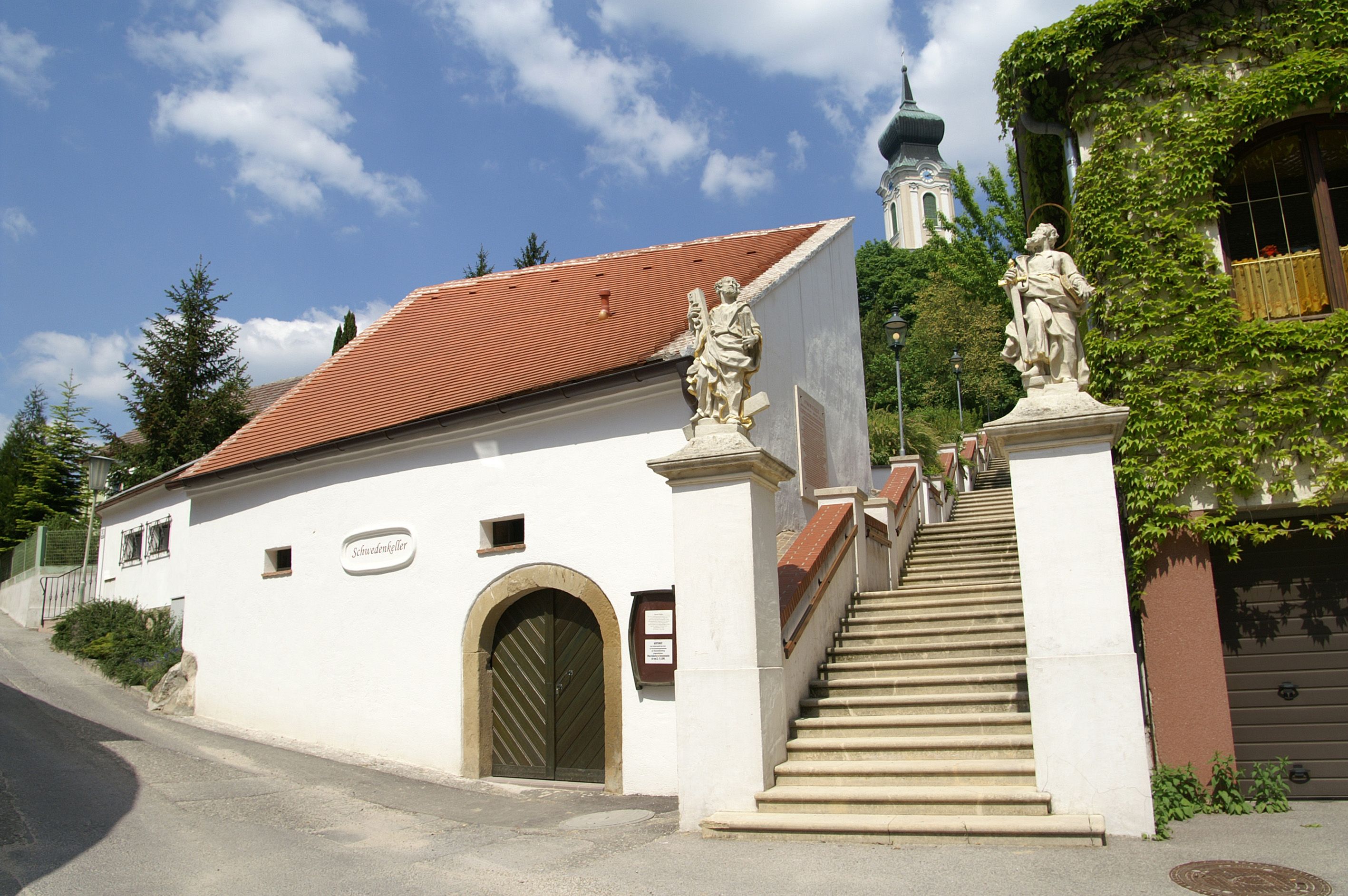 White building with red roof and staircase with statues, church in the background.