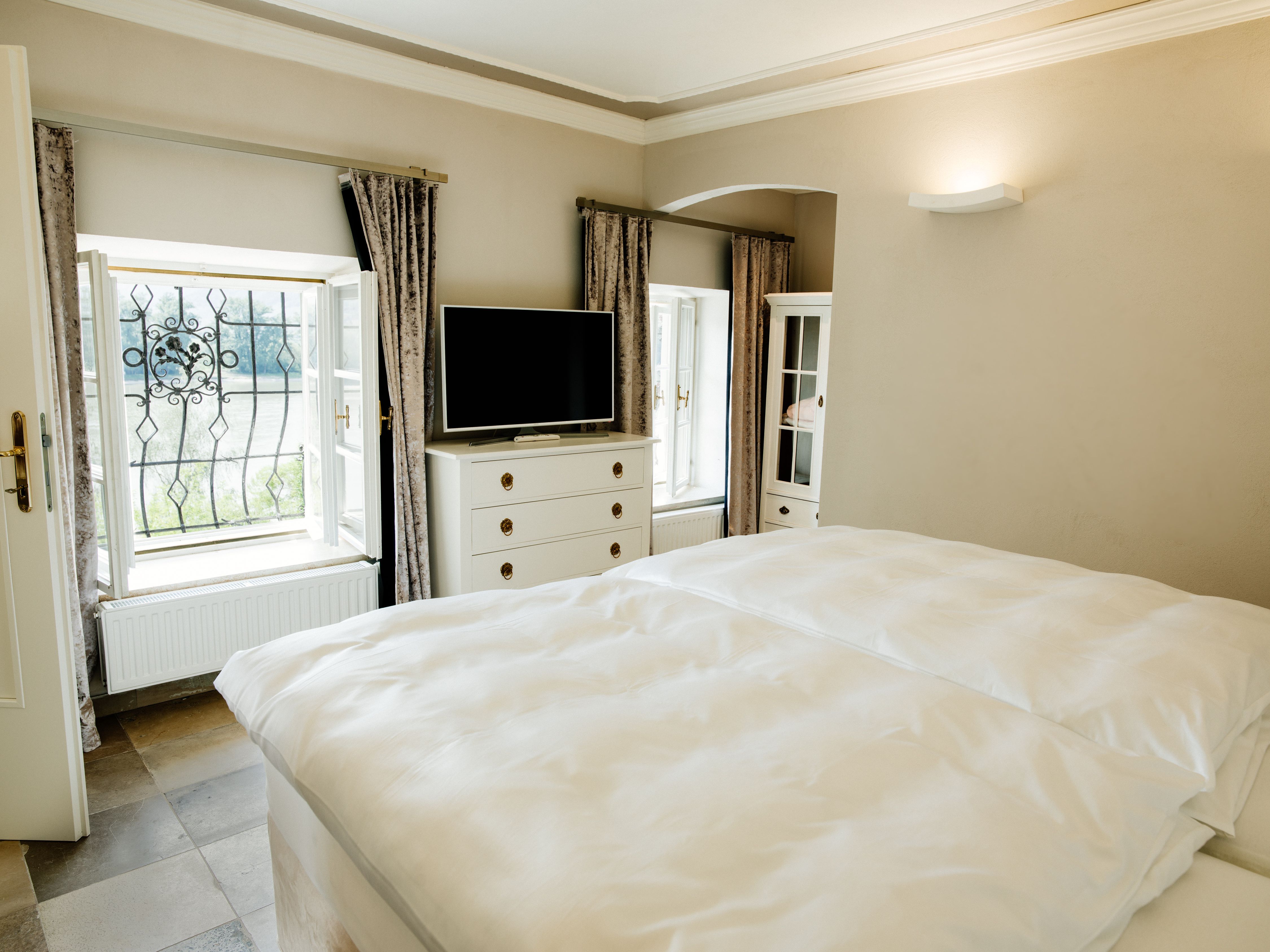 Bright bedroom with double bed, TV, chest of drawers and two windows with curtains.