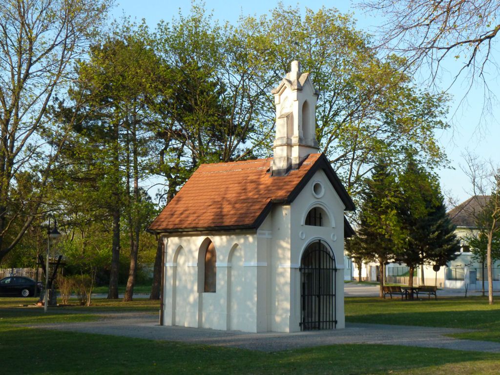 A small white chapel with a red roof in a park surrounded by trees.