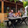 Two men sit at a wooden table outside and taste wine. Surrounded by plants and a wooden building in the background.
