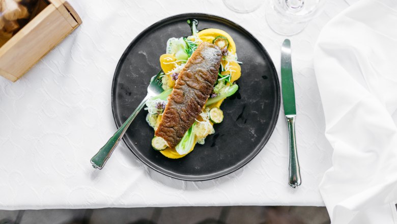 An artfully prepared char fillet on a black plate with vegetables and sauce, on a white table.