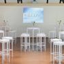 Seminar room with white bar tables and stools, vases of flowers and a screen with the inscription 'Welcome'.