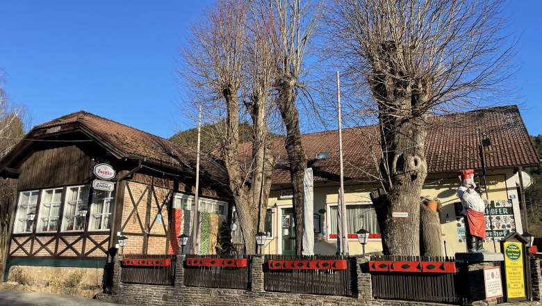 Half-timbered building with red roof tiles, surrounded by bare trees and a fence. A figure dressed as a cook stands in front of the building.