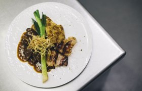 A plate with roast beef, potato gratin, leek and crispy onions on a metal surface.