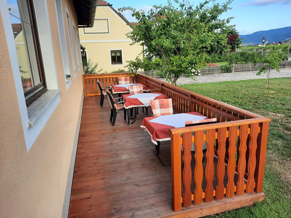 Wooden terrace with covered tables and chairs, surrounded by green landscape.