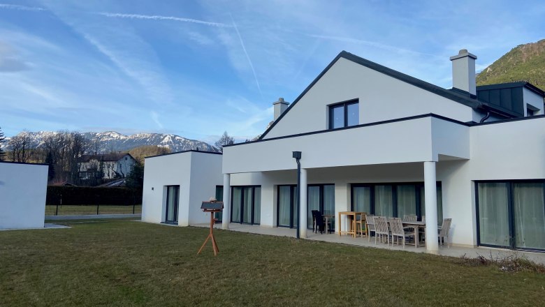 Modern white house with terrace and garden, snow-capped mountains in the background.