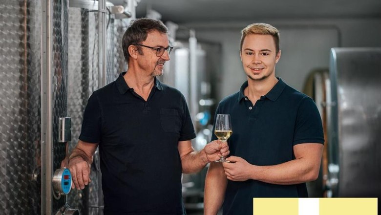 Two men in a wine cellar, one holding a glass of white wine, the other serving a tank.
