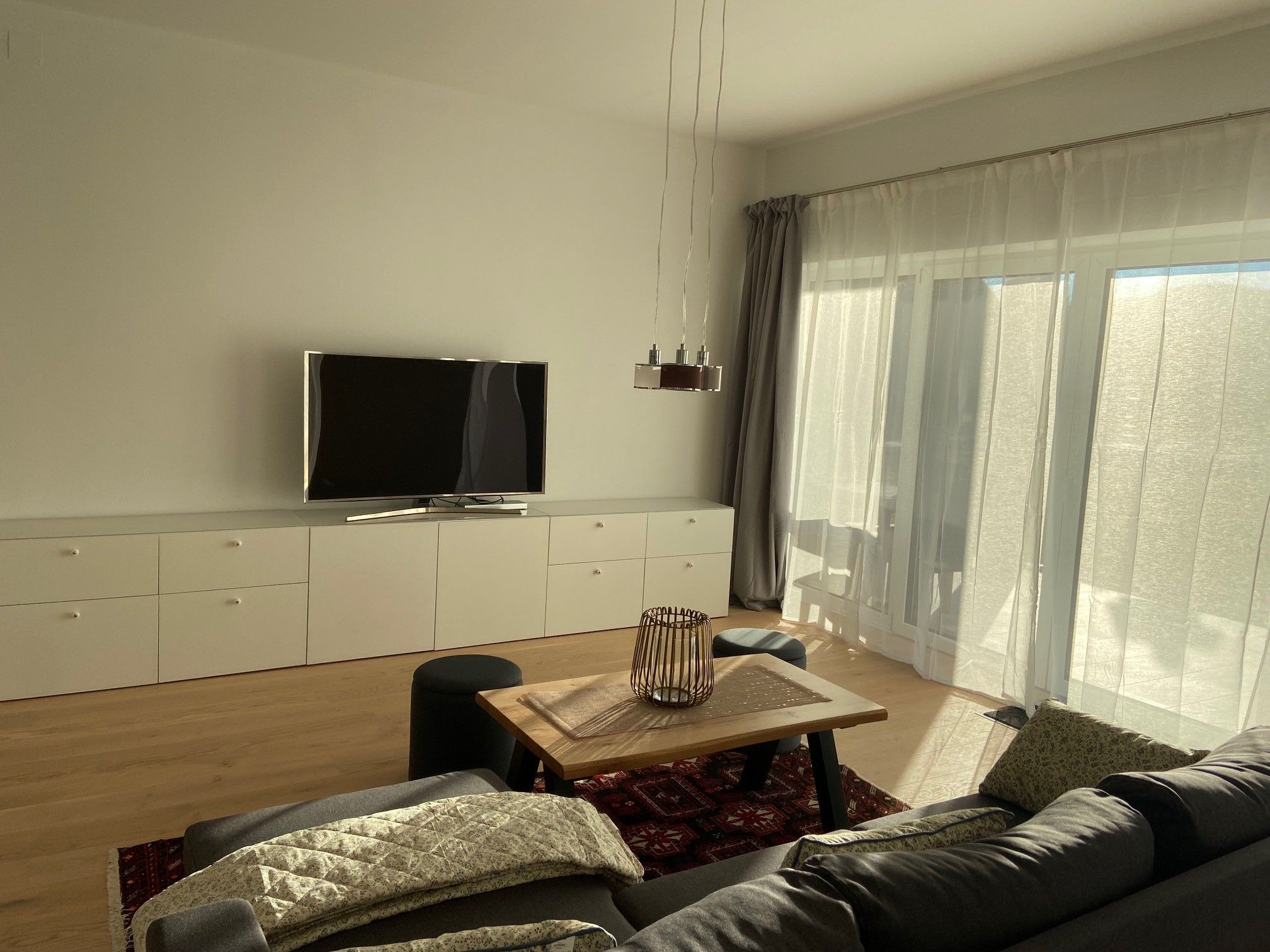 Modern living area with gray sofa, wooden table, carpet, TV and large windows with curtains.