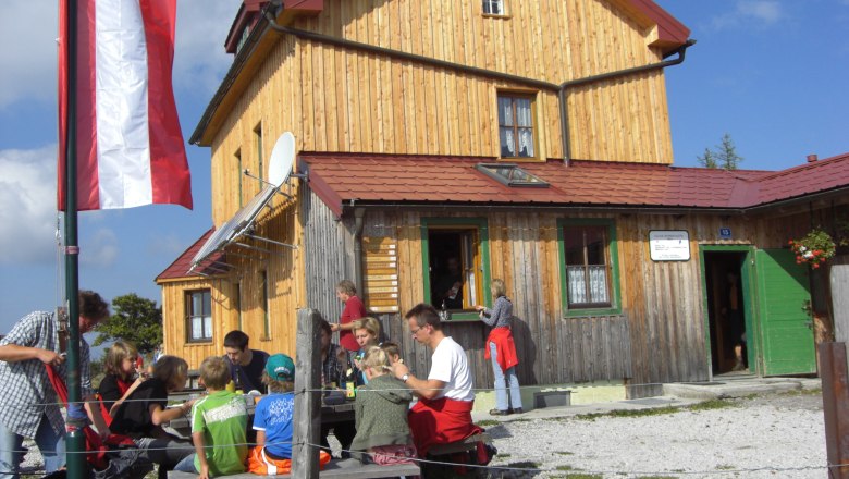 A group of people sit in front of a mountain hut with a wooden fa&ccedil;ade and Austrian flag.