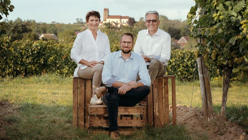 Three people sitting on wooden crates in a vineyard, with a church in the background.