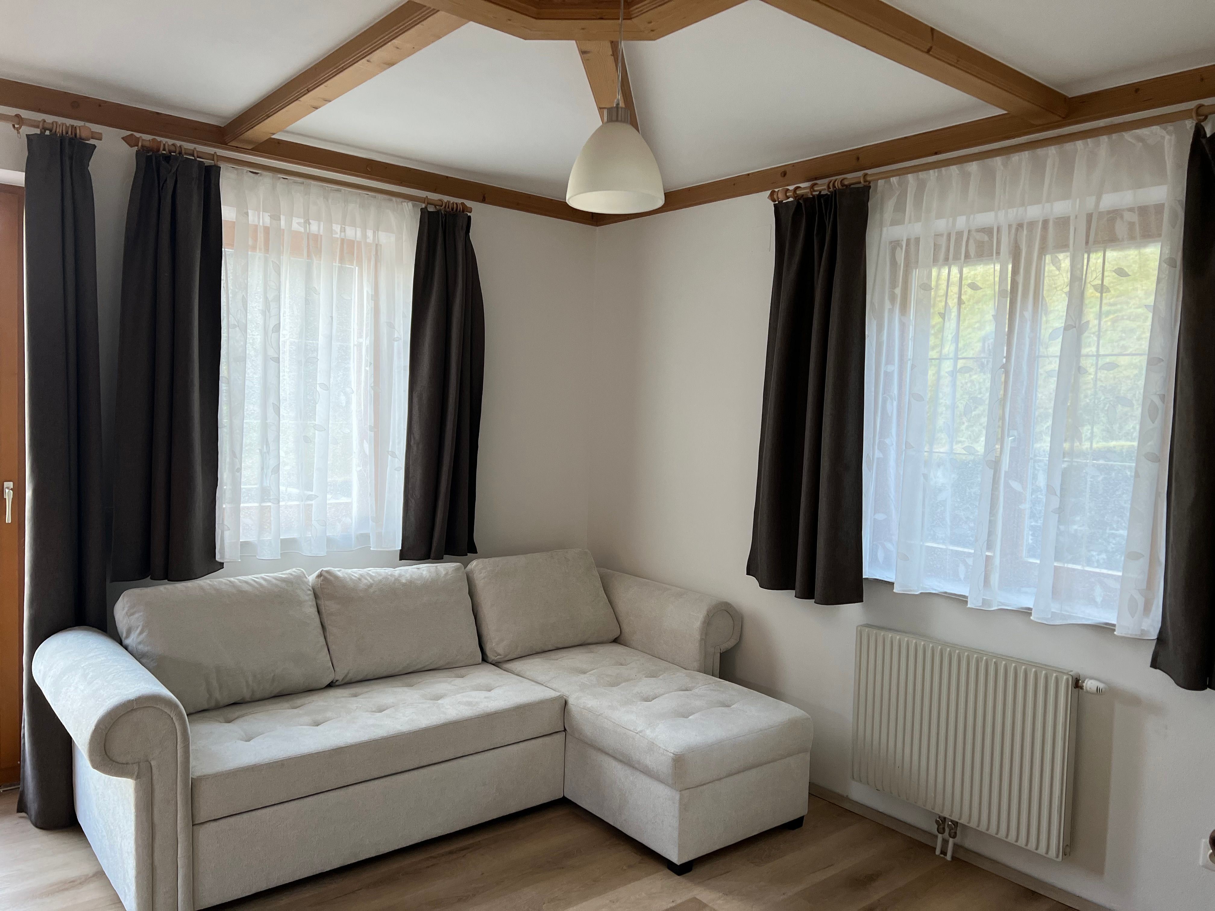 A living room with a beige corner sofa, two windows with dark curtains and a radiator underneath.