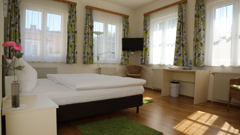 Bright room with bed, desk, TV and several windows with curtains.