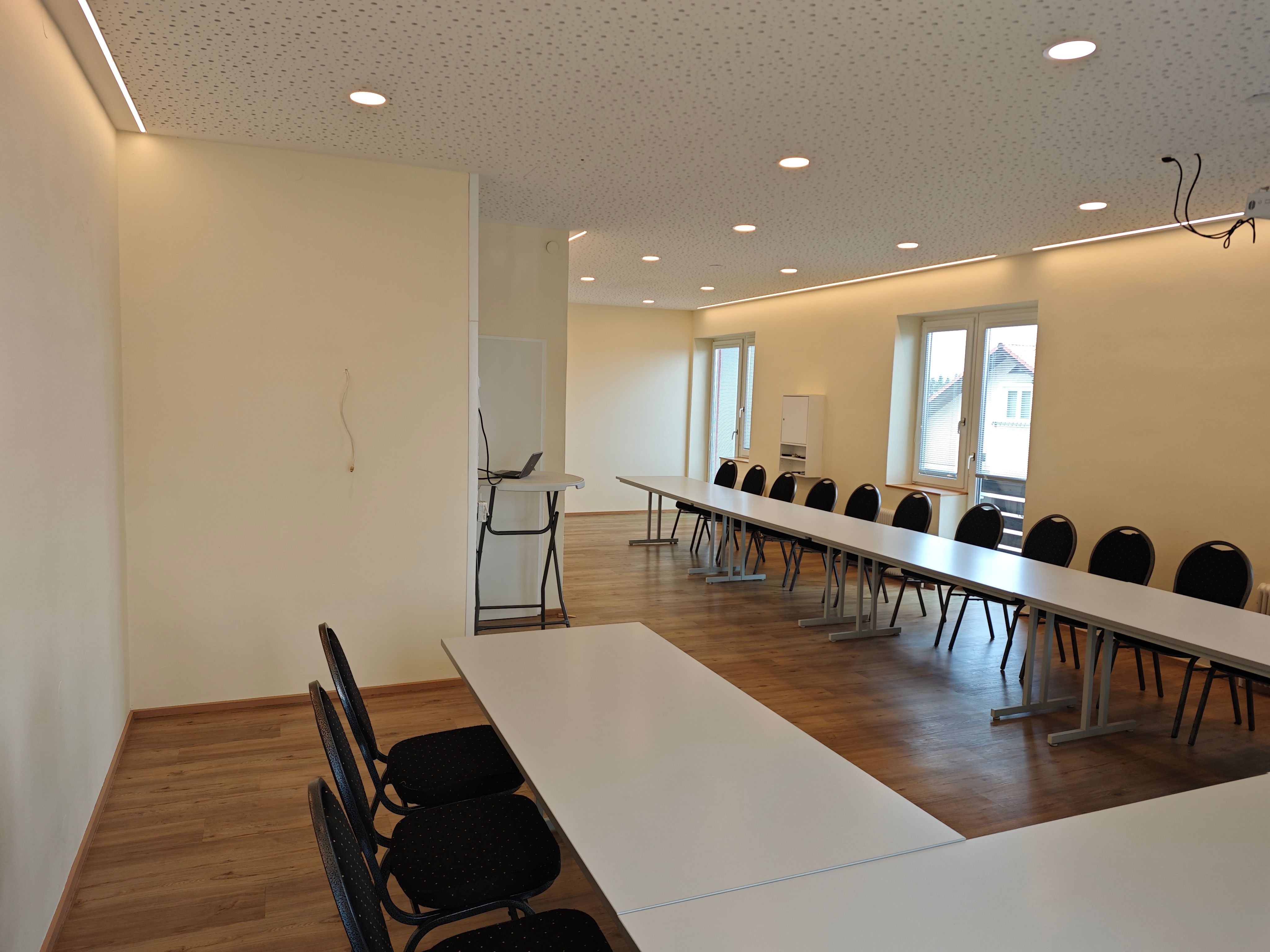 An empty seminar room with long tables and chairs, light-colored wooden floors, and large windows.