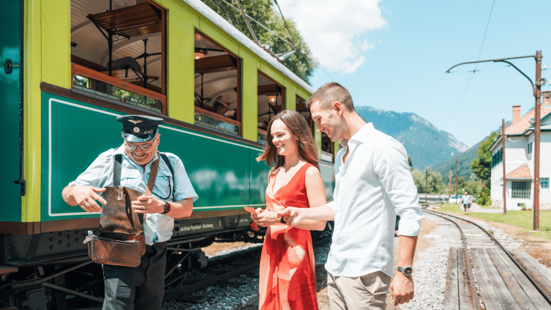 A conductor in uniform shows a couple in front of a green train something from his pocket. Tracks and mountains can be seen in the background.