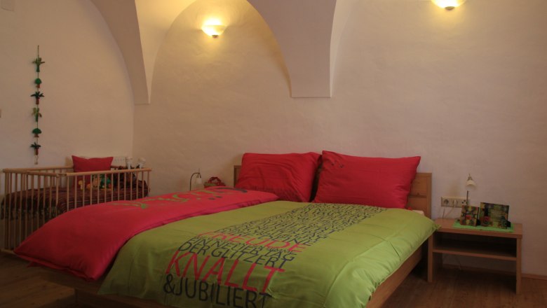 A cosy guest room with a double bed, colorful comforter covers and a baby crib in the background. The walls are white and curved.