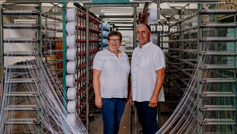 Two people are standing in front of machines with spools of thread in a textile factory.