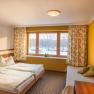 A cozy double room with two beds, yellow walls, a window with curtains, a TV on the wall and a table lamp.