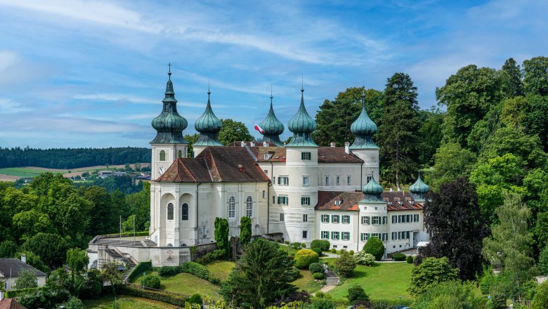 A white castle with several onion domes, surrounded by green countryside and trees, under a blue sky.