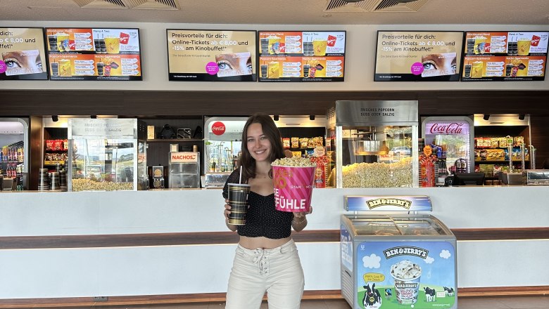 A woman stands in a movie theater foyer in front of a Star Movie sign with popcorn and a drink in her hands.