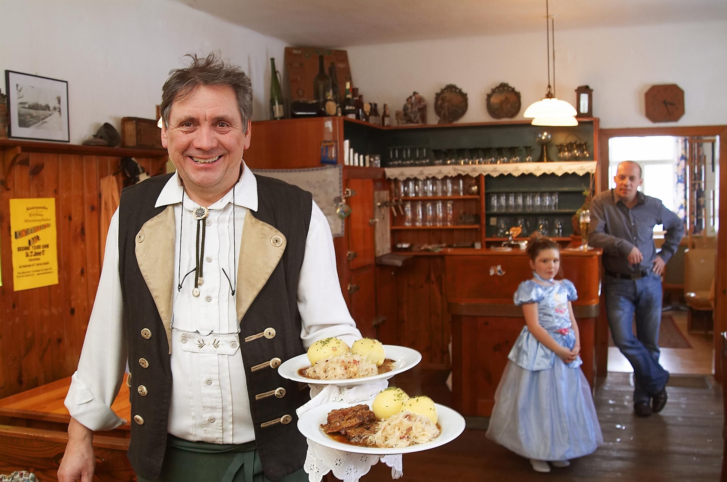 A man in traditional dress serves roast pork with dumplings in a rustic inn. In the background a man and a girl in a dress.