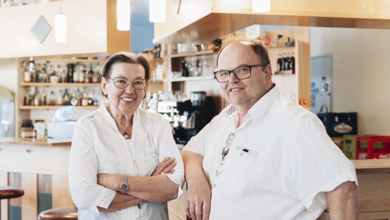 Two people in a café or bar, smiling, in front of a counter with bottles in the background.