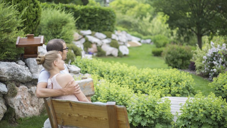 A couple is sitting on a wooden bench in a green garden with bushes and trees.