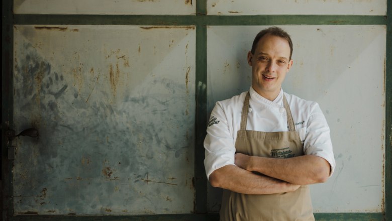 Cook in white uniform in front of rusty metal door, smiling into the camera.